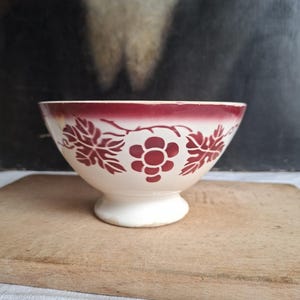 Könnte beinhalten: Eine cremefarbene Keramikschale mit einem dunkelroten Rand und einem sich wiederholenden Muster aus stilisierten roten Blumen und Blättern an der Außenseite. Die Schale hat einen runden Körper, der sich zu einem runden Sockelfuß verjüngt.