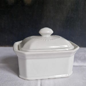 Pode incluir: Uma manteigueira de cerâmica branca com tampa. O prato é retangular com uma base e laterais ligeiramente curvas. A tampa tem um pequeno botão redondo e uma borda decorativa com contas. O fundo é uma imagem escura e desfocada.