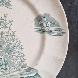 May include: Off-white ceramic plate with a teal transferware design. The design features a rider on a horse jumping over a log, a dog, trees, and water. The plate has a slightly worn appearance.