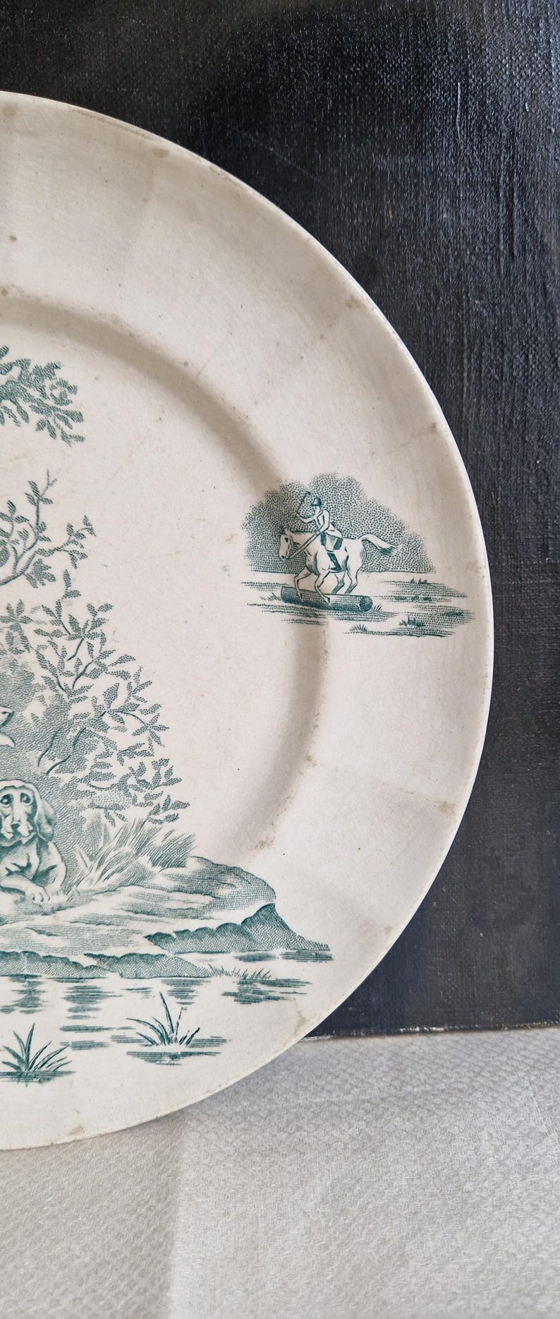 May include: Off-white ceramic plate with a teal transferware design. The design features a rider on a horse jumping over a log, a dog, trees, and water. The plate has a slightly worn appearance.