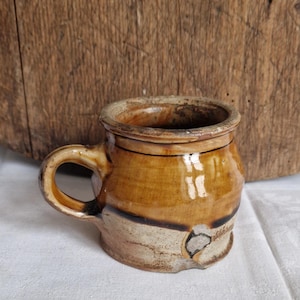 May include: A small, rustic ceramic mug with a handle. The mug features a two-tone design with a light beige base and a warm brown upper section. The mug has a slightly irregular shape, suggesting it is handmade.