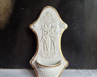 May include: A white porcelain holy water font featuring a gold-trimmed edge and a relief sculpture depicting a religious scene with figures near a cross. The piece includes a small, semi-circular basin at the bottom with vertical fluting.