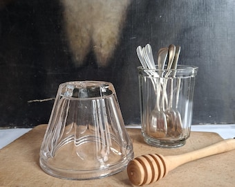 May include: A clear glass bowl turned upside down, a clear glass tumbler containing several small silver-toned metal spoons, and a light-coloured wooden honey dipper. The items are arranged together on a flat, rectangular wooden surface.