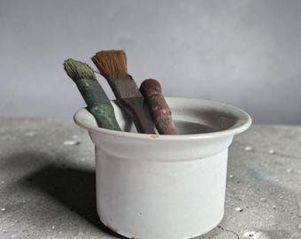 May include: A white ceramic crock with a wide rim containing three paint brushes with wooden handles and worn bristles. The brushes vary in size and colour, with one featuring green paint residue on the handle and bristles.