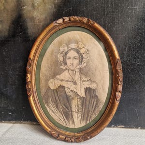 Può includere: Ritratto ovale di donna in cornice dorata. L'immagine in seppia raffigura una donna con un cappello ornato di fiori e un abito con colletto alto. La cornice ha un design ornato e intagliato e un passe-partout verde.