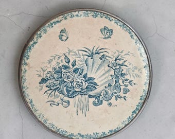 May include: A round decorative plate with a cream-coloured surface featuring a blue floral and shell motif in the centre. Two small butterflies are depicted above the flowers. The plate has a blue floral border and a metallic rim.