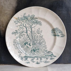 May include: Cream-coloured ceramic plate featuring a teal illustration of two dogs near a tree and a small scene of a person on a log. The plate has a scalloped edge and a vintage aesthetic.