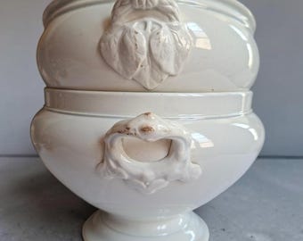 May include: A cream-coloured ceramic tureen with a matching lid, featuring a rounded body and a pedestal base. The lid is adorned with a sculpted leaf-shaped handle, while the base has two ornate, curved side handles with subtle brown distressing.