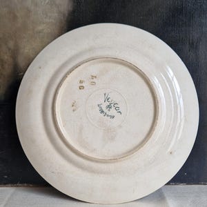 May include: Off-white ceramic plate with a slightly cracked glaze. The underside features the manufacturer's mark "Vedeau Longchamp" in green, along with the numbers "50" and "07". The plate has a simple, classic design.