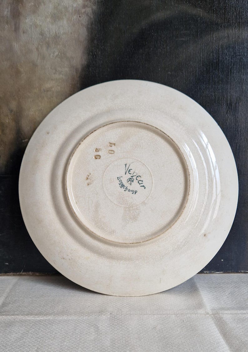 May include: Off-white ceramic plate with a slightly cracked glaze. The underside features the manufacturer's mark "Vedeau Longchamp" in green, along with the numbers "50" and "07". The plate has a simple, classic design.