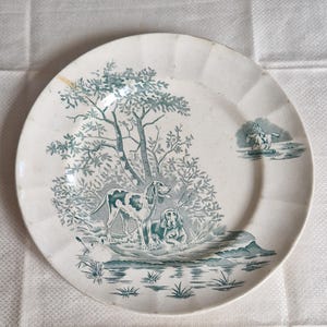 May include: A white ceramic plate with a teal hunting scene. The plate features two dogs near a body of water and a tree. A smaller scene on the right shows a person on a horse. The plate has a scalloped edge.