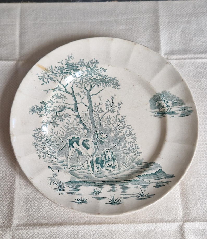 May include: A white ceramic plate with a teal hunting scene. The plate features two dogs near a body of water and a tree. A smaller scene on the right shows a person on a horse. The plate has a scalloped edge.