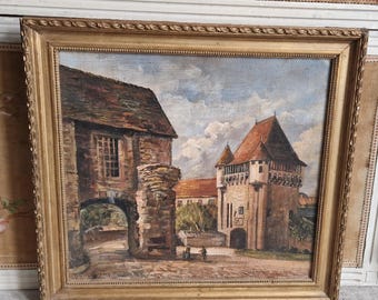 May include: Oil painting of a European town scene in a gold ornate frame. The painting depicts stone buildings with red tile roofs, an arched gateway, and two figures walking on a cobblestone street under a cloudy sky.