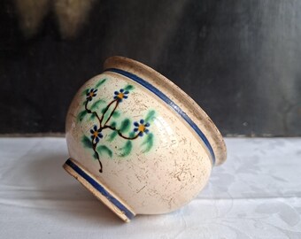 May include: A small, round ceramic bowl with a cream-coloured base featuring a speckled texture. The bowl is decorated with a hand-painted floral motif of blue flowers with yellow centres and green leaves, accented by two horizontal dark blue bands.