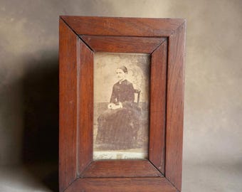 May include: A rectangular wooden picture frame with a dark brown finish containing a sepia-toned photograph of a person seated on a chair. The bottom of the photograph features printed text that reads Fitch, Groton.