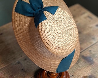 May include: A light brown woven straw hat featuring a flat crown and a wide brim. The hat is decorated with a dark teal ribbon that wraps around the base of the crown and is tied into a bow at the front centre of the hat's crown.