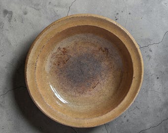 May include: A round ceramic bowl with a wide, flat rim. The bowl features a glossy, honey-coloured glaze with a visible crackle pattern throughout. The centre of the bowl transitions into a darker, brownish-grey tone with fine, dark lines.