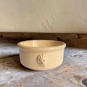 Puede incluir: Un cuenco pequeño y redondo de cerámica de color crema con un acabado liso y brillante. El exterior presenta un relieve moldeado de una cabeza de león centrado en el lateral. El cuenco tiene un borde sencillo y grueso y un diseño abierto.