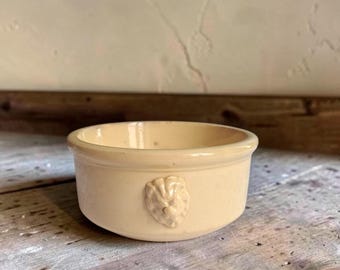 May include: A small, round, cream-coloured ceramic bowl with a smooth, glossy finish. The exterior features a single, raised, moulded relief of a lion's head centred on the side. The bowl has a simple, thick rim and a shallow, open interior design.