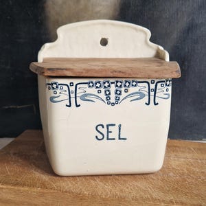 May include: Cream ceramic salt box with a wooden lid. The box has blue decorative patterns and the word "SEL" in blue. The box is mounted on a wooden board.