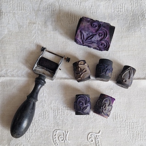 May include: A set of vintage printing rollers with intricate floral designs. The rollers are made of wood and have a dark purple color. The set includes a hand-crank roller and several smaller rollers.