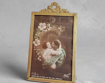 May include: A rectangular brass picture frame with an ornate top crest featuring a bow and floral motif. The frame holds a vintage-style postcard depicting a couple embracing, surrounded by a floral arch, with the text Laissons, dans un baiser, nos deux âmes s'étreindre.