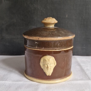 Puede incluir: Un tarro de cerámica marrón con tapa, con un relieve de rostro color crema en la parte delantera. El tarro tiene forma redonda con un borde y base marrón claro. La tapa tiene un pequeño pomo en la parte superior.