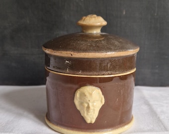 May include: A brown ceramic jar with a lid, featuring a cream-colored face relief on the front. The jar has a round shape with a light brown rim and base. The lid has a small knob on top.