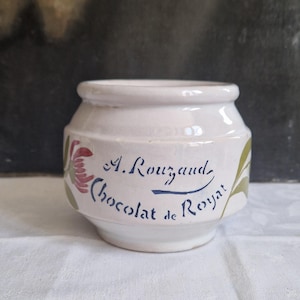 May include: A small, off-white ceramic jar with a rounded shape. The jar features floral and leaf designs in red and green, with the text "A. Rouzaud Chocolat de Royal" in blue script. The jar is likely vintage.