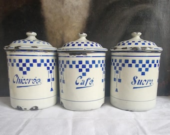 May include: Three vintage white enamel canisters with blue checkered patterns and lids. Each canister is labeled in blue script: "Chicoree", "Cafe", and "Sucre". The canisters have a distressed, aged appearance.