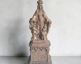 May include: A bronze-toned statue of a crowned figure standing with arms outstretched, flanked by two smaller figures at the base. The statue rests on a rectangular pedestal featuring an embossed architectural relief and the text PIO X PON MAX.
