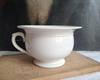 May include: A white ceramic chamber pot featuring a wide, flared rim, a rounded body, and a single curved handle on the side. The vessel sits on a circular pedestal base and has a smooth, glossy finish throughout its entire exterior surface.