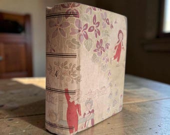 May include: A hardback book with a light beige cover featuring a repeating pattern of purple flowers and green leaves. The spine and front cover display illustrations of figures in red clothing, a tree, and the number 4 printed in black ink.