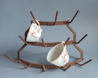 May include: A curved, multi-tiered metal rack with a rusted finish, featuring several upward-pointing prongs. Two white ceramic teacups with delicate pink floral patterns and scalloped edges are hanging from the metal prongs of the rack.