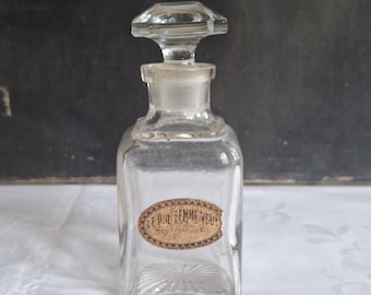 May include: A clear glass perfume bottle with a square base and a faceted stopper. The bottle features an oval-shaped paper label on the front with the text "CE QUE FEMME VEUT" printed in black ink, surrounded by a decorative brown border.