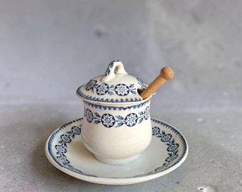 May include: A cream-coloured ceramic honey pot with a matching lid and saucer, decorated with a blue floral pattern. The pot features a small handle on the lid and includes a light brown wooden honey dipper resting inside the container.