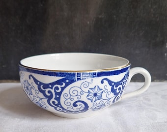 Puede incluir: Taza de té blanca con un diseño floral y de remolinos azul. La taza tiene un borde dorado y un asa. El diseño incluye flores estilizadas y volutas. La taza está sobre una superficie blanca.