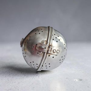 Op de afbeelding: Een bolvormige theezeef van metaal met een geborstelde zilveren afwerking. Het object heeft een scharnierend ontwerp met een klein sluitmechanisme aan de zijkant en talloze kleine ronde gaatjes verspreid over het oppervlak voor losse thee.
