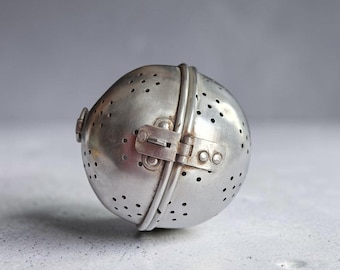May include: A spherical metal tea infuser with a brushed silver finish. The object features a hinged design with a small latch mechanism on the side and numerous small circular perforations distributed across the surface for steeping loose tea leaves.
