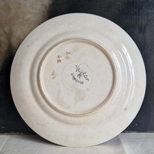 May include: Off-white ceramic plate with a slightly raised rim. The underside features the text "Veneur Longchamp" in a circular design, along with the numbers "50" and "40". The plate has a vintage appearance.