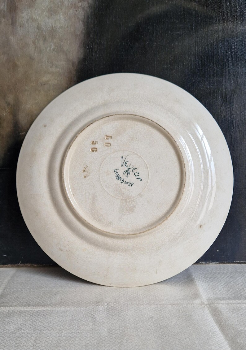 May include: Off-white ceramic plate with a slightly raised rim. The underside features the text "Veneur Longchamp" in a circular design, along with the numbers "50" and "40". The plate has a vintage appearance.