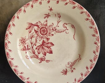 May include: Cream-colored plate with a red floral design. The plate features a central flower with large leaves and a decorative border of smaller floral elements. A vintage-style decorative plate.