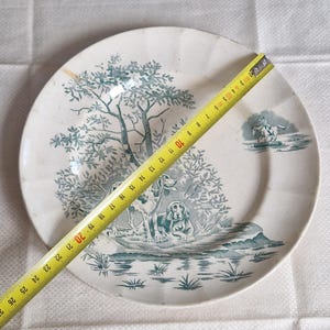 May include: A white ceramic plate with a teal landscape design featuring two dogs, trees, and a figure on horseback. The plate measures approximately 10 inches in diameter.