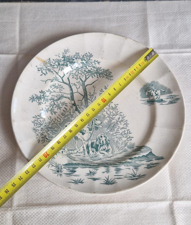 May include: A white ceramic plate with a teal landscape design featuring two dogs, trees, and a figure on horseback. The plate measures approximately 10 inches in diameter.