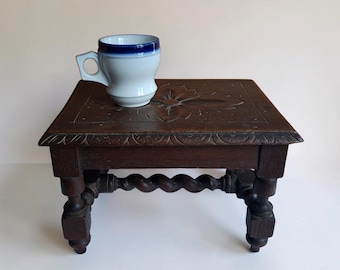 May include: A small, dark brown wooden stool with a carved floral pattern on the top and barley-twist legs, supporting a white ceramic mug featuring a thick, dark blue band around the rim and a geometric, angular handle.
