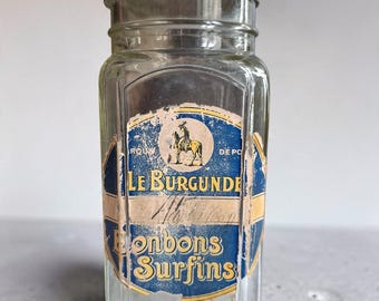 May include: A clear glass jar with a metallic lid and a weathered blue and yellow paper label. The label features an illustration of a horse and rider, with the text LE BURGUNDE and Bonbons Surfins printed in a decorative yellow font.
