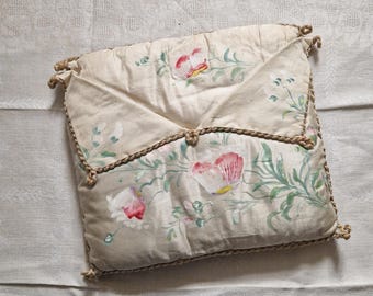 Könnte beinhalten: Cremefarbene, gesteppte Clutch im Umschlagstil mit handgemaltem rosa und rotem Blumendesign. Die Kanten sind mit einem gedrehten Seildetail und geknoteten Akzenten versehen. Die Clutch befindet sich auf einer weißen, strukturierten Oberfläche.