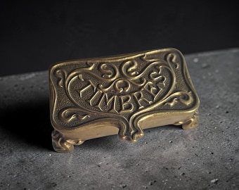 May include: A rectangular brass box with four small feet, featuring an ornate embossed border and the word TIMBRES centred on the lid. The metal surface has a textured, aged finish with raised lettering and decorative scrollwork details.