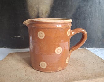May include: A cylindrical ceramic creamer jug with a glossy brown glaze and a repeating pattern of light beige circular spots. The jug features a small pouring spout on the left side and a curved handle on the right side, with a smooth rim.