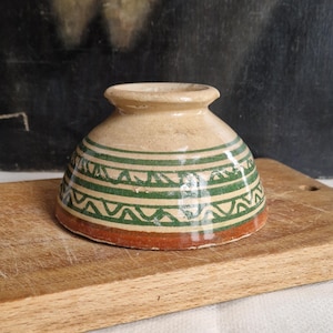 Op de afbeelding: Een kleine, antieke keramische kom met een beige basis en een groen en bruin geometrisch patroon. De kom staat op een houten snijplank. De kom heeft een uitlopende rand en een afgeronde body. De diameter is ongeveer 10 cm.